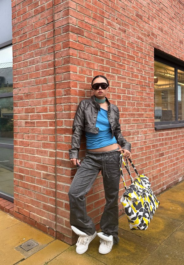 Une personne portant des lunettes de soleil et une tenue streetwear décontractée s’appuie contre un mur de briques en tenant un grand sac cabas à motifs noirs, blancs et jaunes.