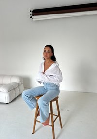 Witte oversized blouse en lichtblauwe flared jeans, zittend op een houten kruk in een minimalistische studio-instelling.