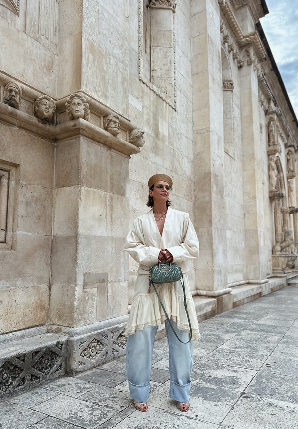 Frau in übergrosser weisser Bluse, weitem Jeans, Sandalen und Hut steht und hält eine kleine grüne Handtasche bei einem verzierten Steingebäude.