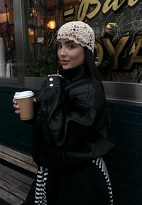 Casaco oversized em pele preta com acabamento texturizado, com botões prateados como detalhe. Modelo segura um copo de cartão castanho e usa um chapéu de croché creme.