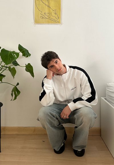 Chaqueta blanca con cremallera y detalles en negro, jeans sueltos de color azul claro, zapatos negros, y una planta verde de fondo contra una pared blanca.