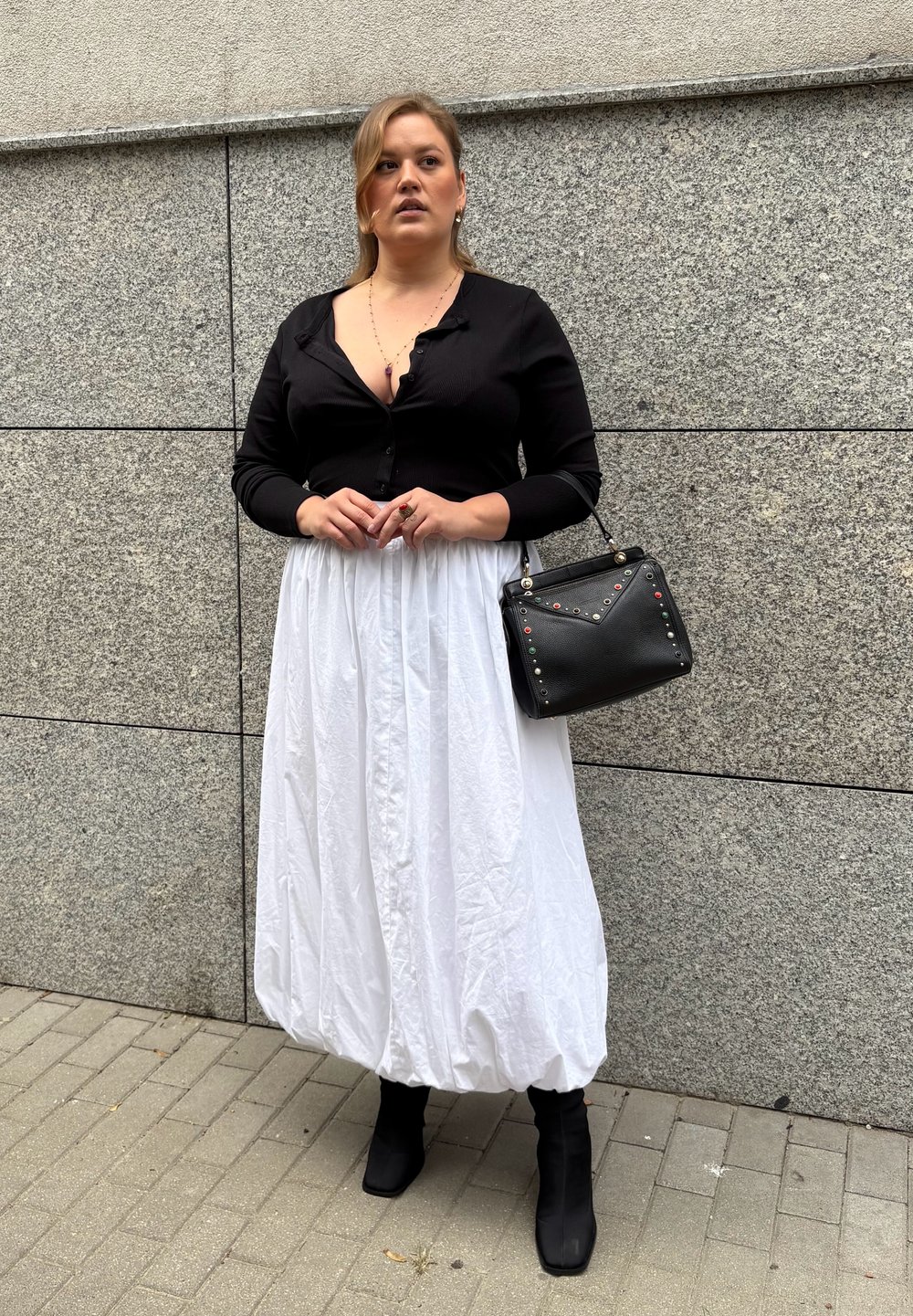 Mujer de pie contra una pared de azulejos grises, vistiendo una blusa negra, una falda globo blanca, botas negras y sosteniendo un bolso negro con tachuelas de colores.