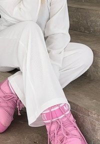 Λευκή ριγωτή μπλούζα με μακριά μανίκια και αντίστοιχα παντελόνια με φαρδιά πόδια σε συνδυασμό με φωτεινές ροζ αθλητικές παπούτσια με κορδόνια και λεπτομέρεια λογότυπου, καθισμένη σε πέτρινα σκαλοπάτια.