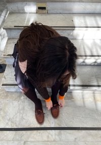 Mocassini in pelle marrone con lacci, indossati su gradini di marmo beige. La persona sta allacciando le scarpe mentre indossa collant scuri e un maglione marrone.
