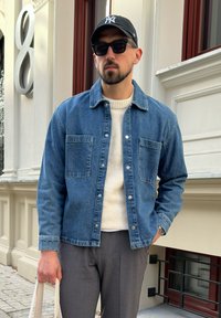Jeansjacka i medelblått med två bröstfickor, lager över en krämfärgad tröja och grå byxor; accessoarer inkluderar en svart keps och solglasögon.