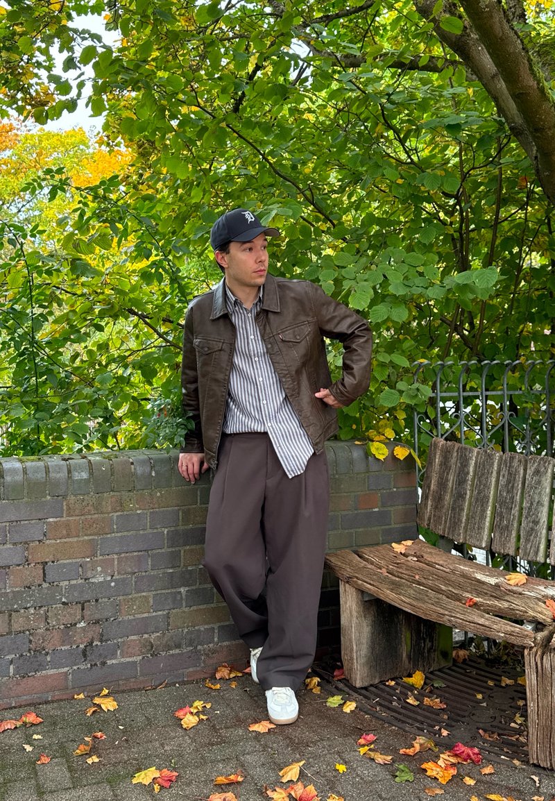 Jeune homme en veste marron et chemise rayée s'appuyant contre un mur en briques près d'un banc en bois, avec des feuilles d'automne sur le sol et des arbres verts en arrière-plan.