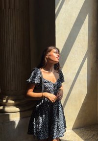 Black floral dress featuring short puff sleeves and a fitted waist. Paired with gold jewellery and a black handbag. Natural light creates shadows.