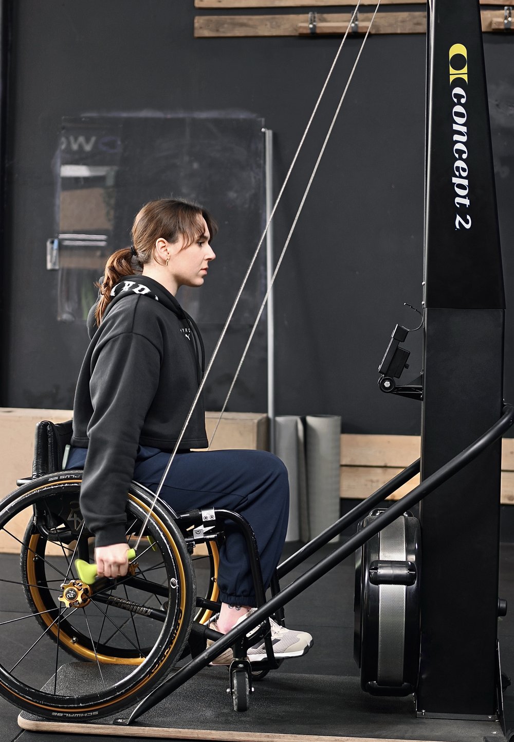 Femme en fauteuil roulant utilisant un rameur Concept2, tirant la poignée avec une expression concentrée dans une salle de sport aux murs sombres.