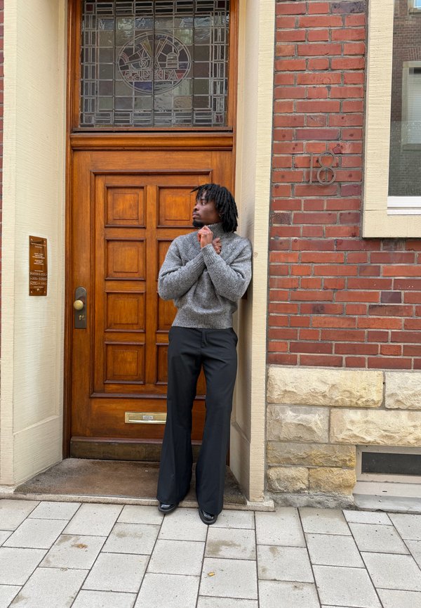 Mężczyzna z czarnymi dredami, ubrany w szary sweter i czarne spodnie, stoi zwrócony w lewo przed drewnianymi drzwiami w budynku z cegły.