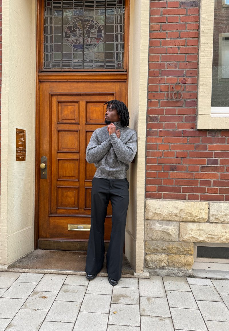 Homme avec des dreadlocks noirs portant un pull gris et un pantalon noir, debout et regardant vers la gauche devant une porte en bois dans un bâtiment en briques.