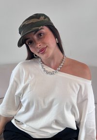 Balta off-shoulder palaidinė, sidabrinių karoliukų vėrinys, kamufliažo kepurė ir dideli sidabriniai žiediniai auskarai. Modelis su ilgu rudais plaukais, sėdintis ant šviesios sofos.