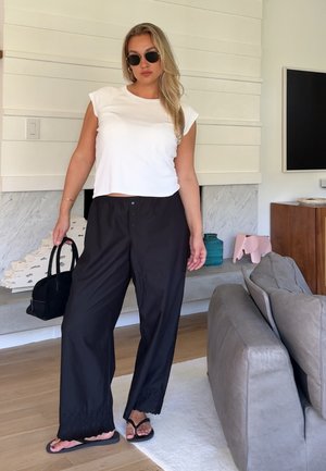 Mujer con gafas de sol, top blanco sin mangas, pantalones anchos negros y chanclas, sosteniendo un bolso negro en el interior.