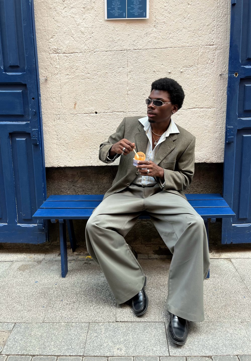 Une personne est assise sur un banc en bois bleu, portant un blazer gris, un large pantalon gris clair et des chaussures noires, tenant une coupe de dessert.