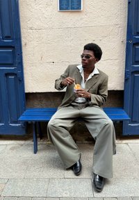 Une personne est assise sur un banc en bois bleu, portant un blazer gris, un large pantalon gris clair et des chaussures noires, tenant une coupe de dessert.