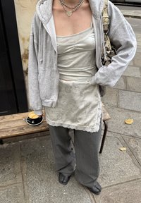 Grå hettegenser, sølvfarget camisole, slip med blondedetaljer, og grå bredbente bukser med nålestriper. Svarte sko, sølvsmykker, og en kaffe på en benk.