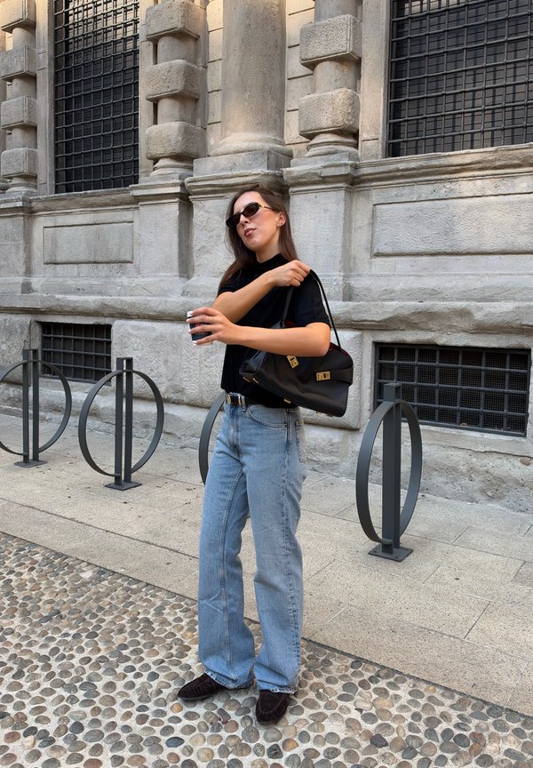 Mujer con gafas de sol, camiseta negra y vaqueros azules, sosteniendo una taza y un bolso negro, de pie en una calle empedrada junto a un edificio de piedra.