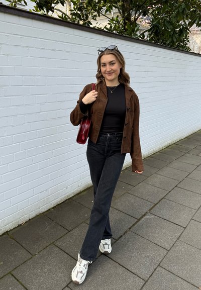 Chaqueta corta de ante marrón sobre una blusa negra, combinada con jeans acampanados negros y zapatillas blancas. Lleva un bolso rojo. Muros blancos al fondo.