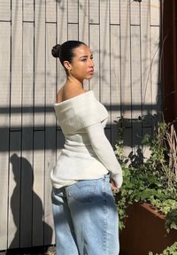 Schulterfreier weißer Pullover mit Strukturstrick, tailliert geschnitten, kombiniert mit hellblauen lässigen Jeans. Der Hintergrund zeigt Holzwände und grünes Pflanzenwerk.