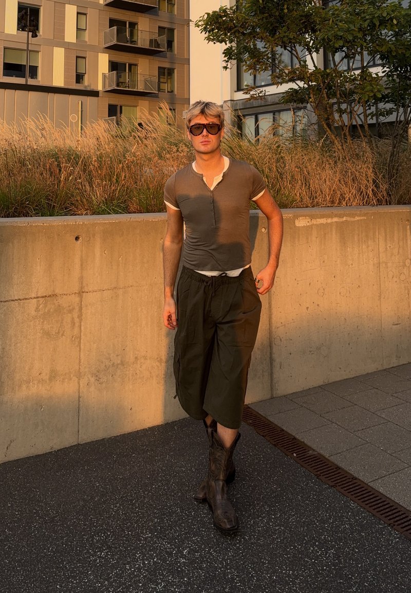 Jeune personne portant des lunettes de soleil, une chemise à manches courtes, un pantalon large et des bottes, marchant sur un trottoir avec des bâtiments et des plantes derrière.