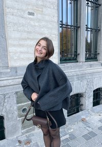 Abrigo de lana gris oscuro con un alto cuello y cierre de botones, combinado con un bolso cruzado marrón y una falda negra. Tela texturizada, ajuste relajado.