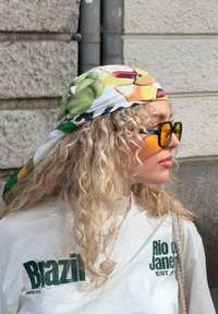 Vit t-shirt med grön text, fruktmönstrad scarf och solglasögon med gulaktig ton. Lockigt hår med en texturerad look. Neutral bakgrund.