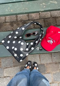 Sac à pois noir et blanc et chaussures, lunettes de soleil écaille, deux bagues, et casquette rouge posés sur un banc vert au-dessus d'un pavé en pierre.