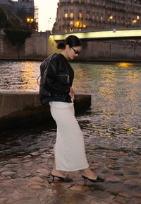 Fekete bőrkabát, fehér kötött maxi szoknya és fekete hegyes orrú magassarkú cipő, amelyet egy nő visel a folyóparton naplementekor, alatta látható kövekkel.