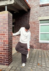 Pessoa de jaqueta branca e calças castanhas a ajustar o sapato enquanto se apoia em um pilar de tijolos fora de um edifício com paredes de tijolos e janelas.