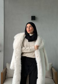 Abrigo largo de piel sintética blanca sobre un suéter beige de canalé, falda negra con estampado y hiyab negro. El entorno tiene un diseño minimalista con una pared gris.