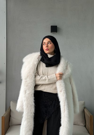 Abrigo largo de piel sintética blanca sobre un suéter beige de canalé, falda negra con estampado y hiyab negro. El entorno tiene un diseño minimalista con una pared gris.