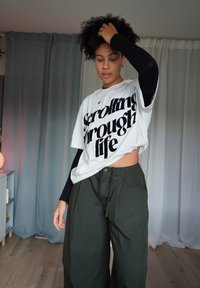 Tricou alb supradimensionat cu text negru îndrăzneț „Scrolling through life”, asortat cu pantaloni largi verde închis. Tricou negru cu mâneci lungi, vizibil dedesubt.