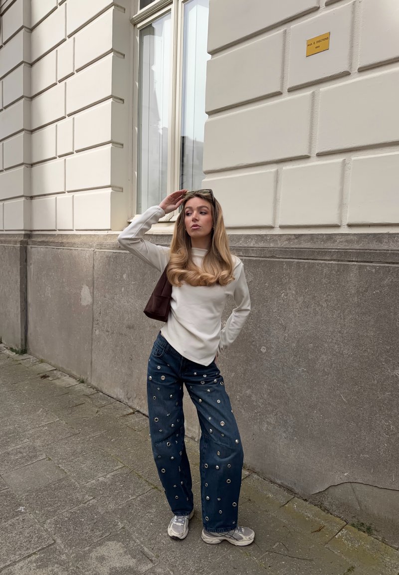 Νέα γυναίκα με λευκό μπλουζάκι και διακοσμημένο τζιν με φαρδιά μπατζάκια στέκεται σε πεζοδρόμιο της πόλης, κρατώντας το ένα χέρι στο κεφάλι και μια καφέ τσάντα στον ώμο.