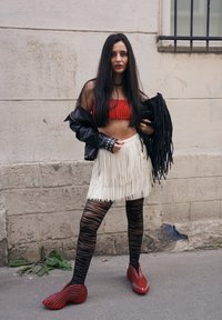 Top corto a righe rosse, gonna bianca frangiata, giacca di pelle nera con borchie, collant a righe nere e scarpe rosse dalla texture. Sfondo urbano.