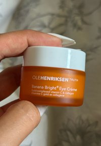 Pomarańczowy szklany słoik z białą pokrywką, oznaczony napisem "OLEHENRIKSEN Truth Banana Bright+ Eye Crème", z złotym tekstem i kremowymi detalami.