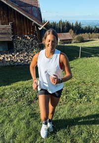 Femeie care alergă pe un deal acoperit cu iarbă, aproape de un morman de lemne și pădure, sub un cer senin în timpul zilei.