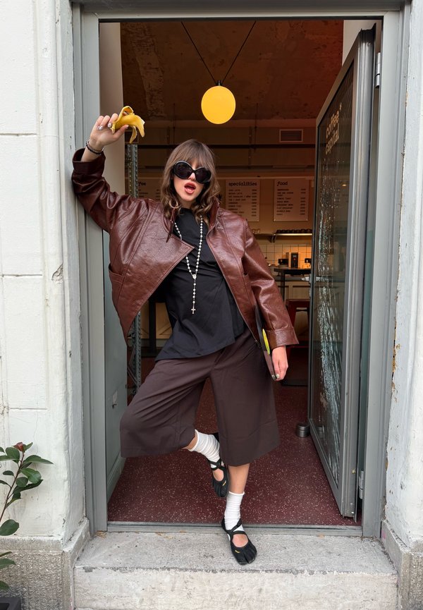 Mujer con gafas de sol y chaqueta de cuero marrón que sostiene un plátano pelado, posando juguetonamente en la entrada de una cafetería.