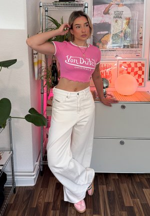 Camiseta rosa corta con texto blanco "Von Dutch Originals", combinada con jeans anchos blancos y zapatillas rosas claras. Atuendo moderno y casual.
