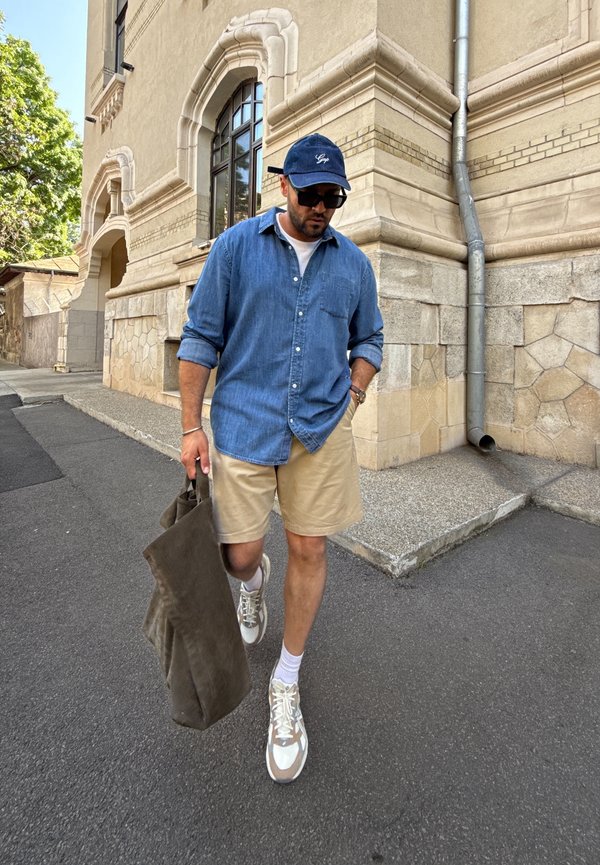 Jeanshemd, beige Shorts, graue Stofftasche und beigen Sneakers. Schwarze Kappe und Sonnenbrille. Geht auf einer gepflasterten Straße in der Nähe eines Steingebäudes.