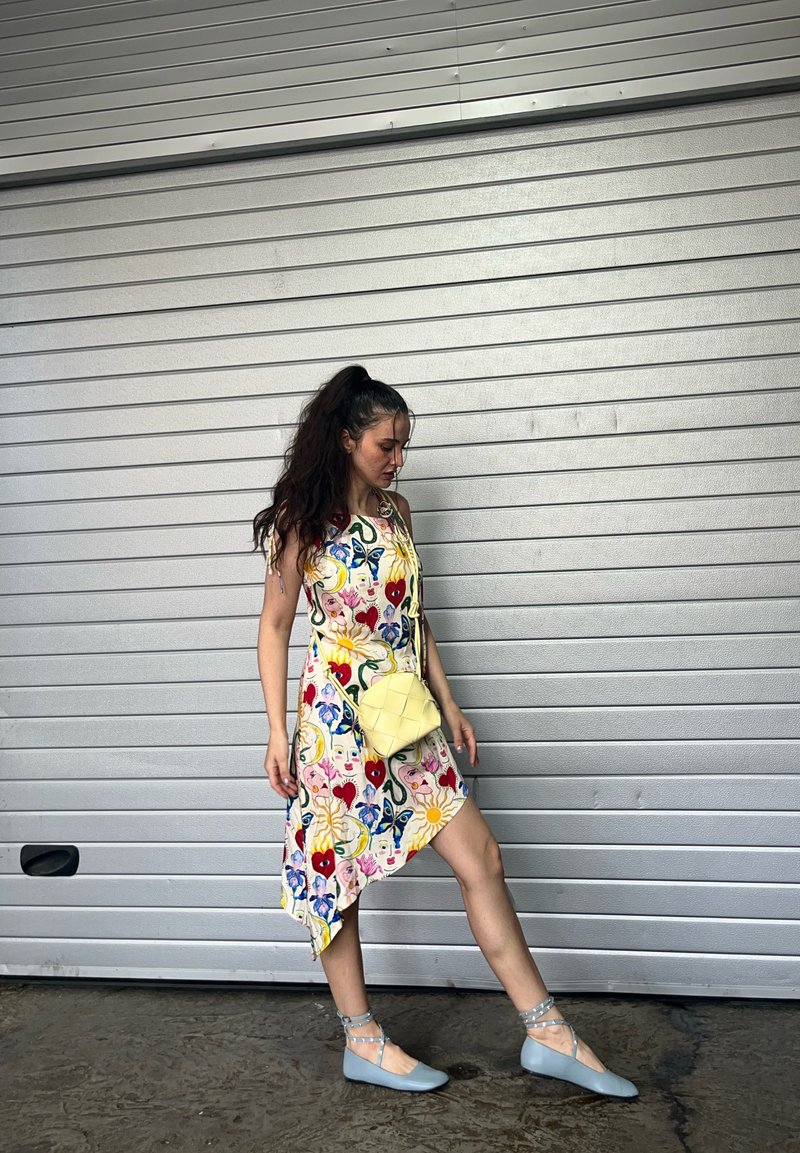 Asymmetrisches Blumenkleid mit bunten Mustern, gelbe Umhängetasche und hellblaue Ballerinas mit Knöchelriemen, vor einem metallischen Hintergrund.