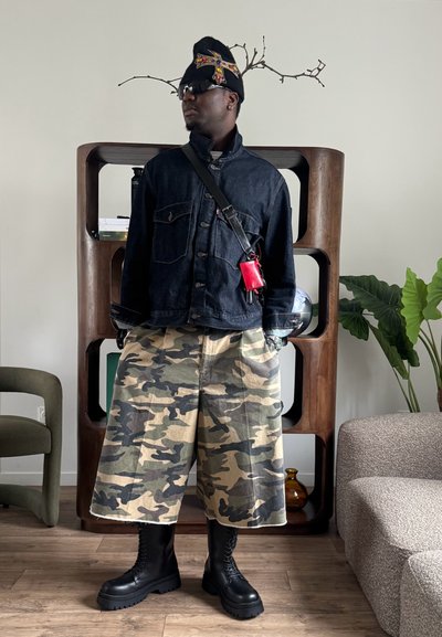 Hombre con chaqueta de mezclilla oscura, pantalones cortos anchos de camuflaje, botas negras con cordones, gorro negro adornado y gafas de sol, de pie en el interior.