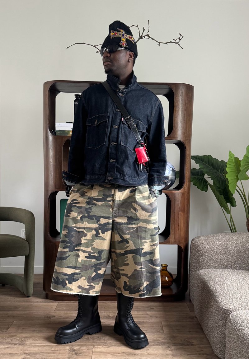 Homme portant une veste en jean sombre, un short large camouflage, des bottes noires à lacets, un bonnet noir orné et des lunettes de soleil, debout à l'intérieur.