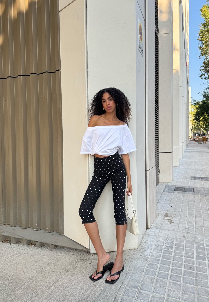 Femme aux cheveux bouclés portant un haut blanc épaules nues, un pantalon capri noir à pois, des sandales noires, tenant un sac à main blanc, appuyée contre un bâtiment.