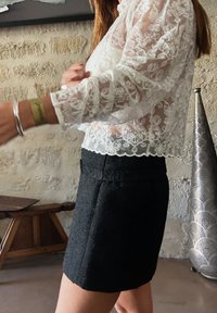 Bela prozorna čipkasta bluzica s cvetnimi vzorci in valovitim robom, v kombinaciji s prilagojeno črno teksturirano mini krilom. Vidni srebrni dodatki.