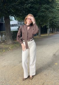 Ruskea pitkähihaine paita kaulakiedolla, vaaleat leveälahkeiset housut ja leopardikuosiset kengät. Seisoo sorapolulla, jota reunustavat puut.