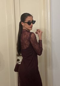 Γυναίκα σε διαφανές μπορντό δαντελένιο φόρεμα, φορώντας μαύρα γυαλιά ηλίου cat-eye, κρατώντας μια μικρή μπορντό τσάντα, στέκεται μπροστά από μια πόρτα.