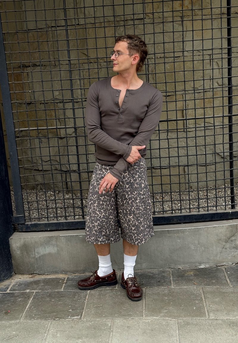 Homme portant des lunettes, un henley marron à manches longues, un short à motifs jusqu'aux genoux, des chaussettes blanches et des chaussures marron, debout devant une clôture en treillis métallique.