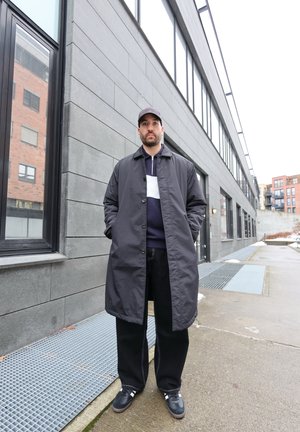 Hombre con un abrigo largo gris oscuro, pantalones negros, zapatillas y una gorra gris de pie en la acera de una ciudad junto a un edificio gris.