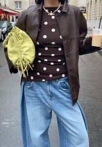 Veste en cuir marron portée sur une chemise marron à pois, associée à un jean large bleu clair et un sac pochette jaune, tenant une boisson glacée.