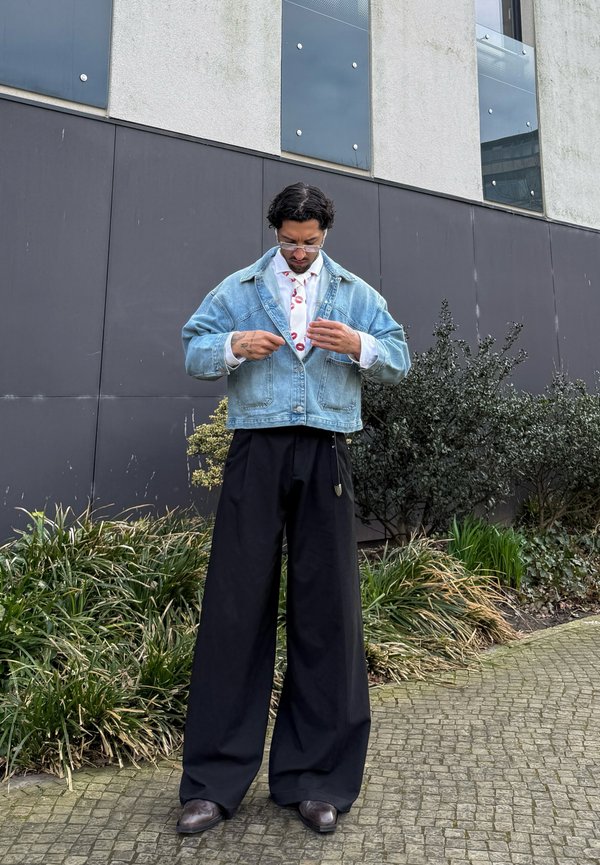 Férfi, aki állítja a szájlenyomatos nyakkendőt, farmerdzsekit, fehér inget, bő, fekete nadrágot és szemüveget visel, kövezett úton áll növények és épület közelében.