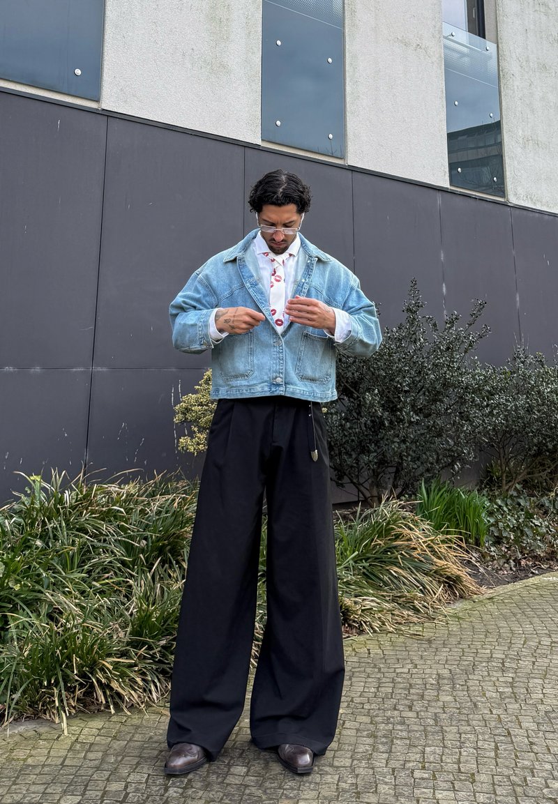 Homme ajustant une cravate à motif empreinte de lèvres, portant une veste en jean, une chemise blanche, un pantalon noir large et des lunettes, debout sur un chemin pavé près des plantes et d’un bâtiment.